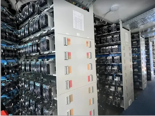 Inside a mining container: rows of ASIC miners on shelves on both sides, with gray breaker panels between them