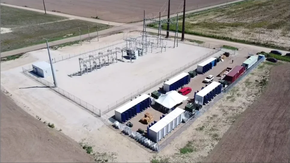 Aerial view of a bitcoin mining site: a fenced electrical substation beside rows of white mining containers on a gravel pad